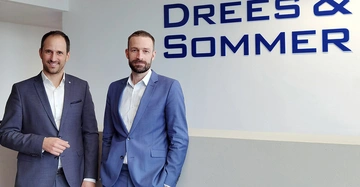 Zwei Männer tragen Anzüge und stehen vor einer Wand. Die Wörter Drees & Sommer sind in Blau auf der Wand zu lesen. Ein Mann lächelt und der andere schaut ernst.