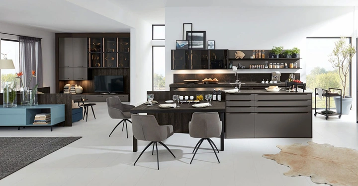 Moderne Küche mit Esstisch aus dunklem Holz gedeckt und grauen Stühlen. Im Hintergrund ein Wohnbereich mit Fernseher und Regalen. Lichtdurchfluteter Raum mit großen Fenstern und Pflanzen.