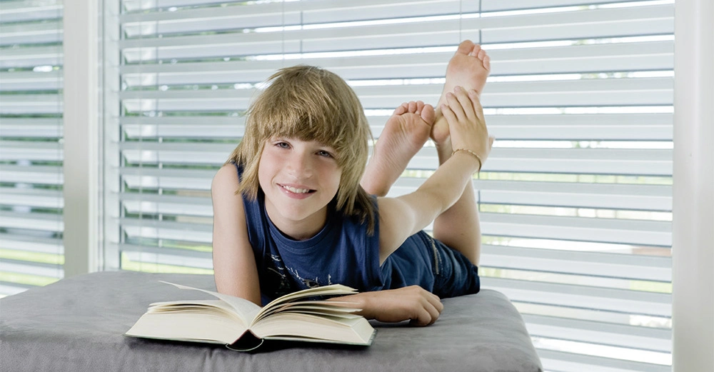 Ein Junge liegt auf einem grauen Sofa liest ein aufgeschlagenes Buch und lächelt während er sich in einem modernen Wohnzimmer mit vertikalen Jalousien befindet.