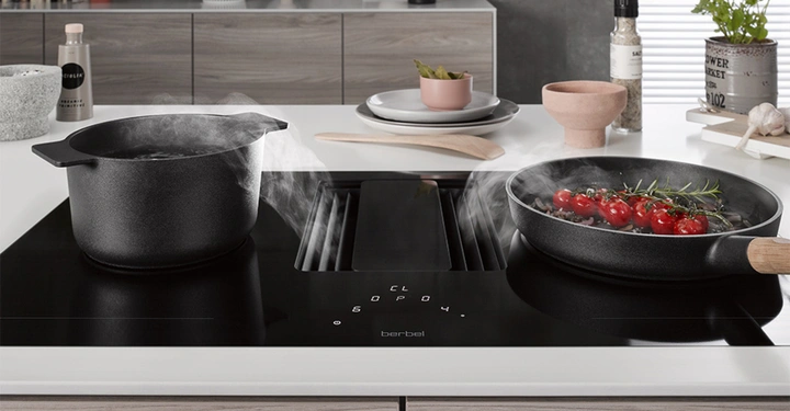 Ein Topf und eine Pfanne mit dampfendem Essen stehen auf einem modernen Herd mit integrierter Dunstabzugshaube in einer modernen Küche. Im Hintergrund sind Gewürze und Küchenutensilien zu sehen. Text im Bild: "berbel"