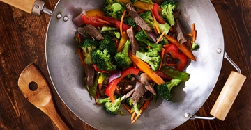 Wok mit Gemüse und Fleischststücken wird erhitzt hölzerner Löffel liegt daneben dunkler Holztisch Hintergrund