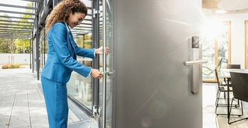 Eine Geschäftsfrau im blauen Anzug öffnet eine Glastür mit einem elektronischen Zutrittssystem (TECTIQ von ABUS) an einem modernen Bürogebäude.