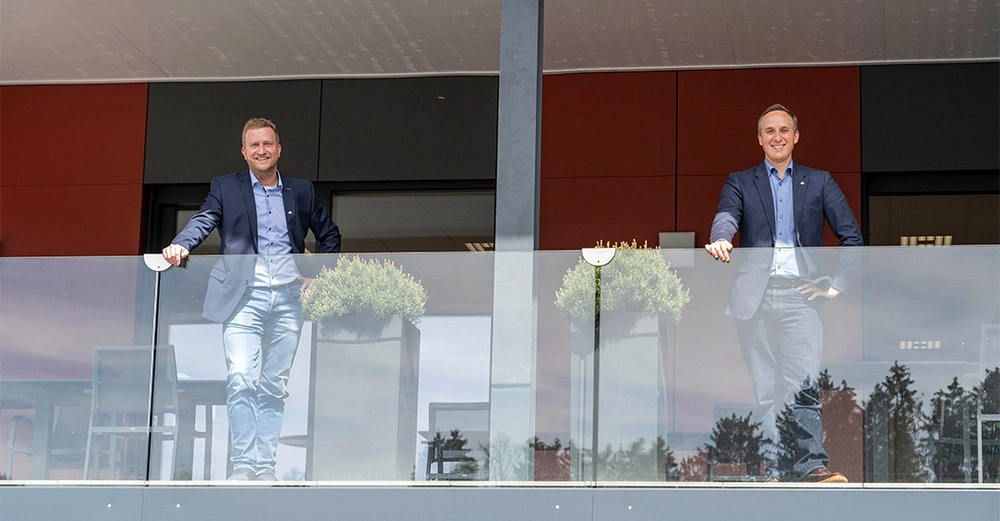 Zwei Männer lehnen sich an einem Glasgeländer auf einem Balkon. Beide tragen blaue Blazer und Jeans. Neben ihnen stehen Pflanzenbehälter. Der Hintergrund zeigt ein modernes Gebäude.