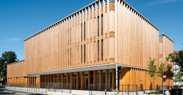 Ein modernes Holzgebäude steht auf einer Straßenecke an einer Kreuzung Tagsüber bei klarem Himmel umgeben von Bäumen und Gehwegen mit einem Fußgängerampel daneben und Zäunen um das Gebäude herum