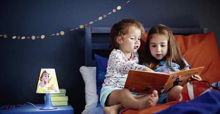 Galerie-Bild 1: Zwei Mädchen lesen in einem Bett ein Buch in heller Bettwäsche ein Kinderzimmerlampe mit Zeichentrickfiguren leuchtet und eine Girlande hängt an der Wand.