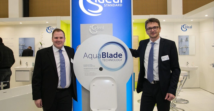 Galerie-Bild 7: Zwei Männer stehen neben einem Toilettenbecken mit der Aufschrift AquaBlade Technology vor einem Ideal Standard Display in einer Badezimmerausstellung.
