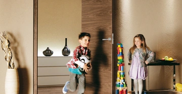 Junge läuft durch offene Tür mit Fußball im Arm, Mädchen steht neben hohem Turm aus Bauklötzen. Im Hintergrund moderne Möbel und Dekorationen in einem gut beleuchteten Raum.