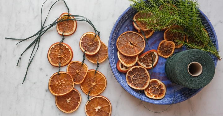 Getrocknete Orangenscheiben sind auf grünem Band aufgereiht auf einer Marmoroberfläche daneben ein blauer Teller mit weiteren Orangenscheiben grünen Zweigen und einer grünen Garnrolle.