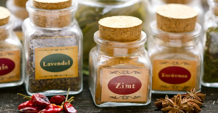 Mehrere Glasgefäße mit Korkdeckeln und Etiketten wie Lavendel Zimt Kardamom und Anis stehen auf einer Holzoberfläche umgeben von getrockneten Chilischoten und Sternanis.