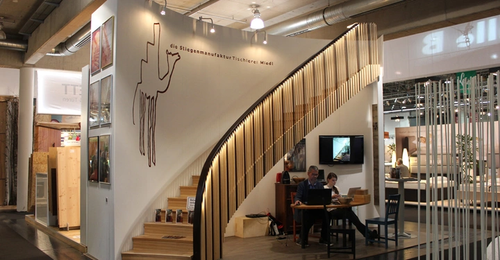 Galerie-Bild 6: Holztreppe ragt elegant in einem offenen Messebereich auf, zwei Personen arbeiten an einem Tisch darunter, umgeben von modernen Möbel- und Architekturinstallationen. Text auf der Wand: die Stiegenmanufaktur Tischlerei Miedl