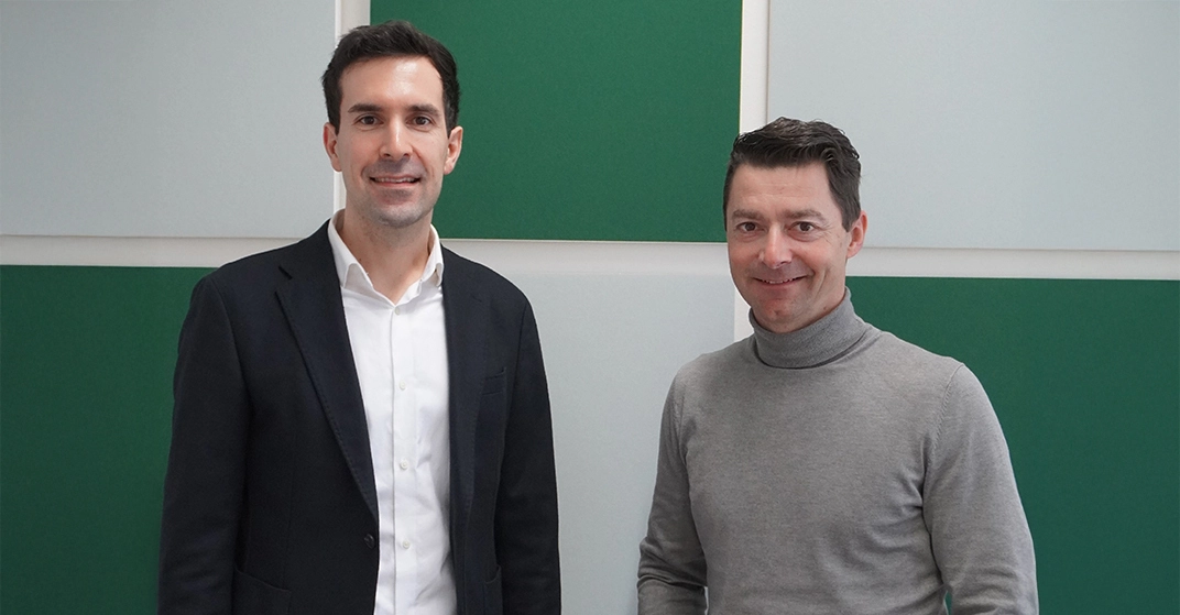 zwei Männer in Businesslook stehen vor grün-weiß gekachelter Wand und lächeln in Kamera