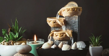 Ein kleiner Brunnen fließt über Steinstufen in einem Raum mit grüner Kerze, Muscheln, Kieselsteinen und Topfpflanzen auf einem dunklen Hintergrund.