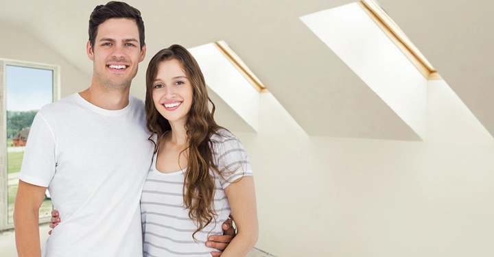 Lächelndes Paar steht umarmend in einem lichtdurchfluteten Raum mit Dachfenstern und weiß gestrichenen Wänden. Im Hintergrund ist ein Fenster mit grüner Landschaft zu sehen.