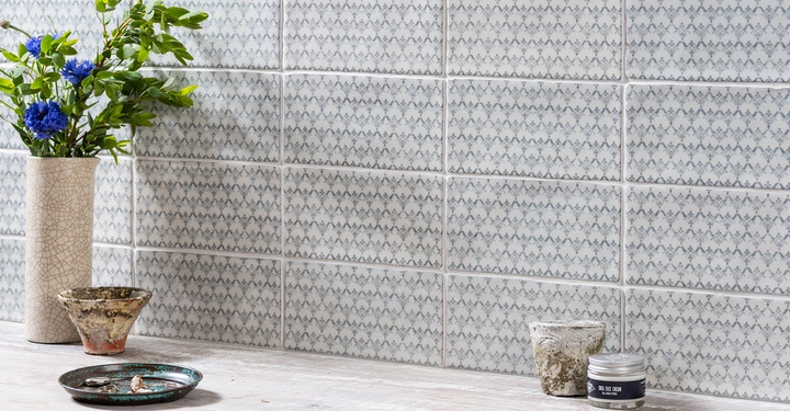 Vase mit blauen Blumen steht auf einem Holztisch neben einer Schale und zwei kleinen Gefäßen vor einer mit gemusterten Fliesen dekorierten Wand.