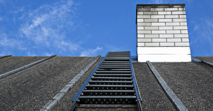 Schwarze Leiter steigt eine raue Dachfläche hinauf ein weißer gemauerter Schornstein ragt aus dem Dach in einem klaren blauen Himmel mit wenigen Wolken empor