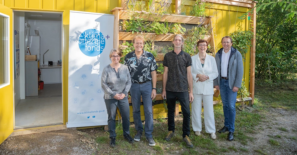 Fünf Personen stehen nebeneinander vor einem vertikalen Garten und einem gelben Gebäude. Ein Banner mit der Aufschrift klima energie fonds www.klimafonds.gv.at steht links neben einer offenen Tür.