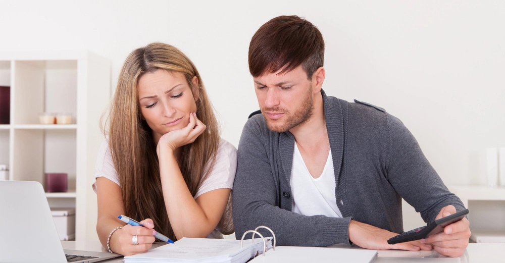 Zwei Personen studieren Dokumente in einem weißen Raum. Die Frau hält einen Stift und sieht besorgt aus, während der Mann einen Taschenrechner benutzt. Ein Laptop steht auf dem Tisch.