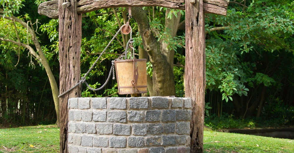 Ein hölzerner Eimer hängt an einer Kette über einem gemauerten Brunnen im Freien umgeben von grünen Bäumen und Gras.