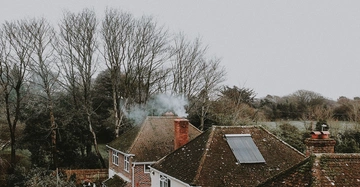 Dach eines Backsteinhauses raucht aus einem Schornstein umgeben von kahlen Bäumen und Wiesen an einem grauen Tag
