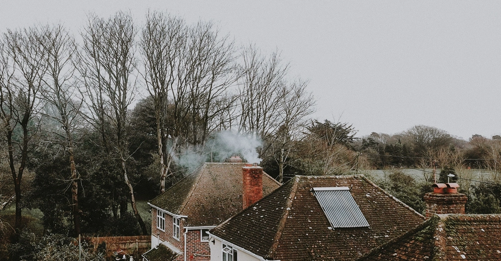 Dach eines Backsteinhauses raucht aus einem Schornstein umgeben von kahlen Bäumen und Wiesen an einem grauen Tag