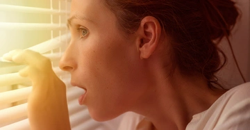 Frau mit braunen Haaren spioniert durch Jalousien durch, ihr Blick wirkt geschockt und überrascht.