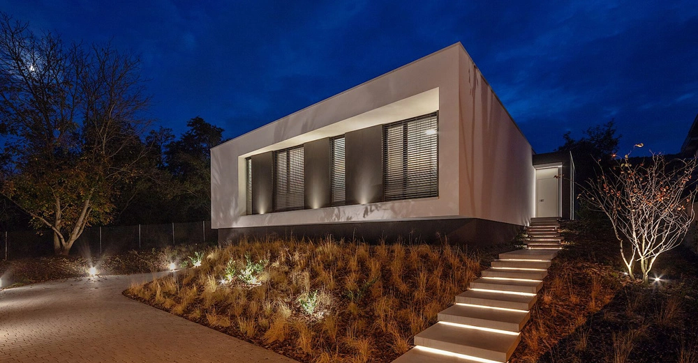 Ein modernes weißes Haus steht auf einem leicht erhöhten Grundstück. Eine beleuchtete Treppe führt zum Eingang. Umgeben von Bäumen und Vegetation am Abendhimmel.
