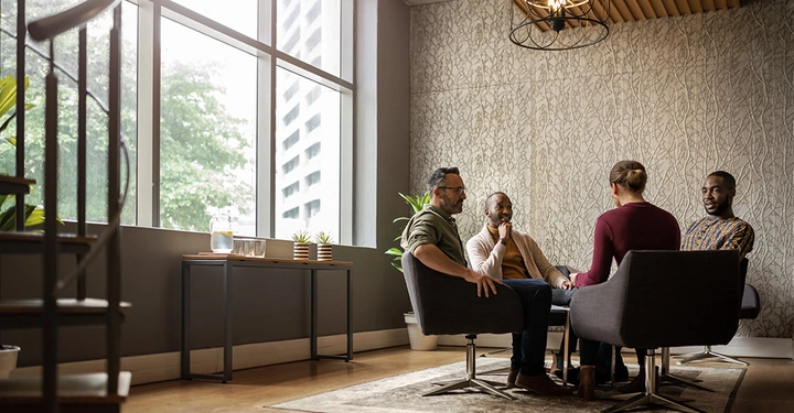 Eine Gruppe von vier Personen sitzt in einem modernen Raum mit großen Fenstern und bespricht etwas miteinander während sie auf bequemen Stühlen um einen kleinen Tisch sitzen.