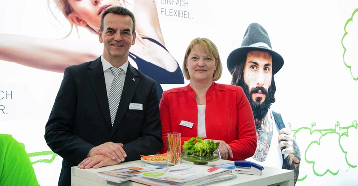 Galerie-Bild 1: Zwei Personen stehen lächelnd an einem Messestand mit Broschüren und Snacks Eine bunte Wand mit Abbildungen von Menschen im Hintergrund Text: Einfach Flexibel und teilweise verdeckt
