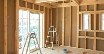 Innenansicht eines Holzhauses in Leichtbausweise. Holzständerwände mit Dämmung, ein großes Fenster an der linken Seite, 2 Leitern im Rauminneren