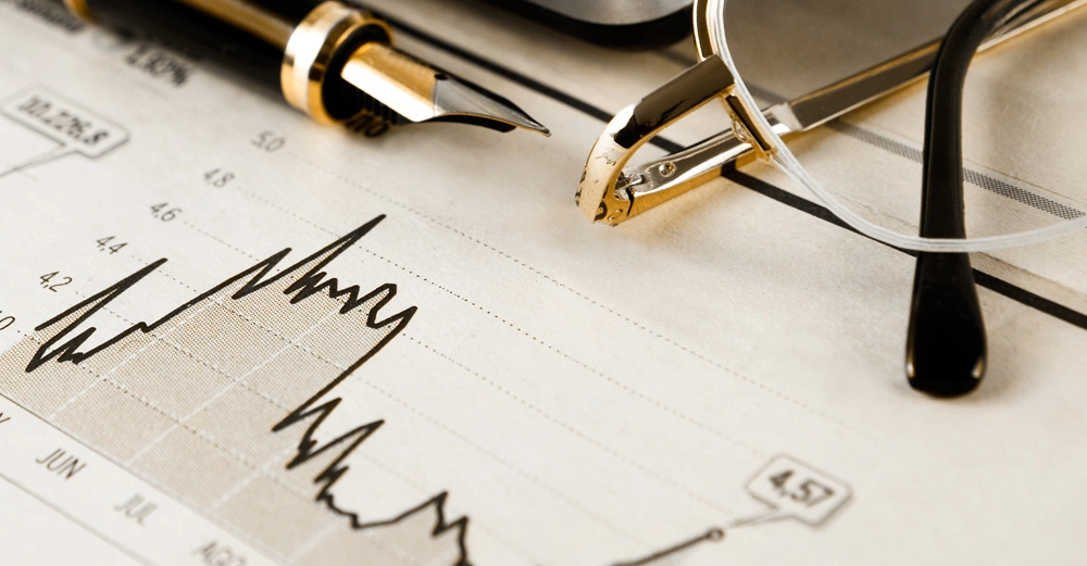 Füllfederhalter liegt auf einem Finanzdokument mit einer Börsenkursgrafik daneben eine Lesebrille auf einem Schreibtisch.