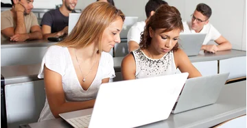 mehrere junge Menschen in sommerlicher Kleidung sitzen vor Laptops in einem Hörsaal.