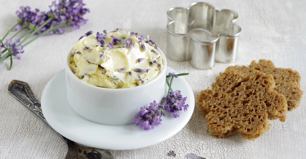 weiße Tasse mit Untertasse voll mit Kräuterbutter, garniert mit lilalen kleinen Lavendelzweigen, daneben blumenförmig ausgestochenes Vollkornbrot auf weißer Tischdecke