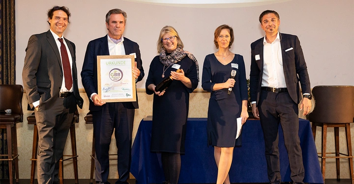 Galerie-Bild 3: Fünf Personen stehen nebeneinander in einem Raum zwei halten eine Urkunde mit der Aufschrift URKUNDE GBB 2021 eine Frau hält ein Mikrofon und spricht.