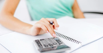 Person tippt auf Taschenrechner auf Schreibtisch im Büro hält Stift in anderer Hand und hat aufgeschlagenes Notizbuch vor sich.