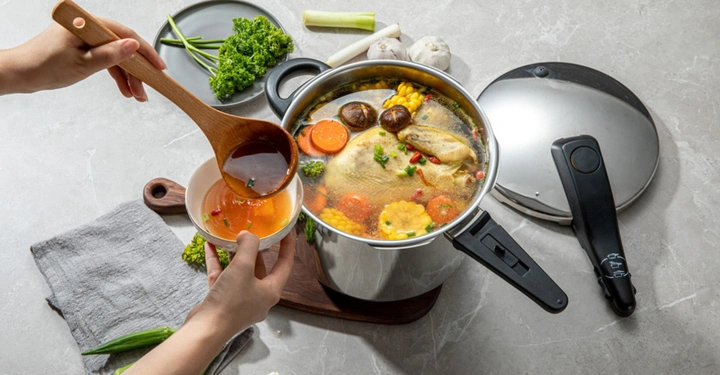 geöffneter Schnellkochtopf (Kelomat) steht auf Tisch und Frau schaufelt den Inhalt, einen üppigen Gemüseeintopf, in eine kleine Essschüssel. 