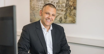 Zehetner in seinem Büro mit dunklem Anzug und leger getragenem Hemd lächelt in Kamera