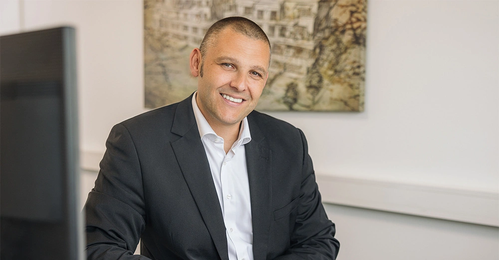 Zehetner in seinem Büro mit dunklem Anzug und leger getragenem Hemd lächelt in Kamera