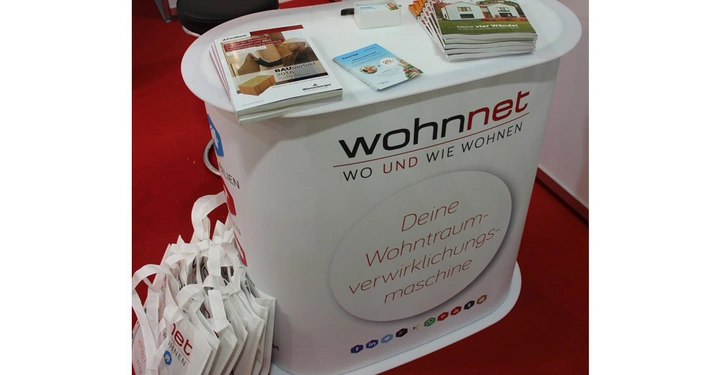 Galerie-Bild 2: Ein weißer Messestandtisch mit der Aufschrift wohnnet WO UND WIE WOHNEN und Deine Wohntraum-verwirklichungs-maschine im Vordergrund auf rotem Teppichboden Zeitschriften und Flyer liegen oben auf dem Tisch.