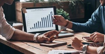 Zwei Personen diskutieren mit Stiften in der Hand über ein Balkendiagramm auf einem Laptopbildschirm an einem Holztisch in einem modernen Büro mit Pflanzen im Hintergrund.