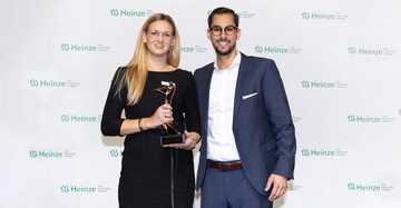 Zwei Personen posieren an einer Wand mit Heinze-Logo der Mann im Anzug die Frau im schwarzen Kleid hält eine Trophäe mit der Aufschrift Heinze ╗ARCHITECT'S DARLING AWARD 2019╗ BALDUS GIGANT GABIONEN