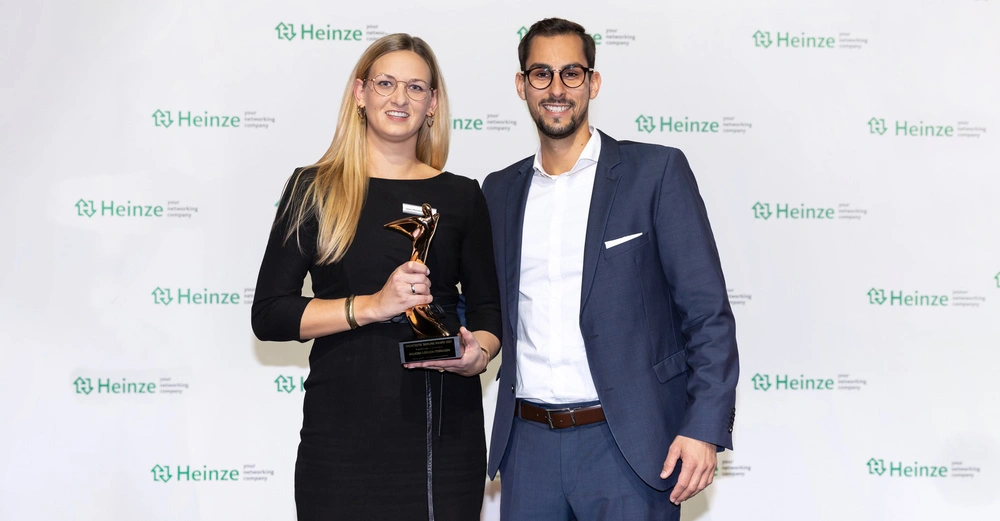 Zwei Personen posieren an einer Wand mit Heinze-Logo der Mann im Anzug die Frau im schwarzen Kleid hält eine Trophäe mit der Aufschrift Heinze ╗ARCHITECT'S DARLING AWARD 2019╗ BALDUS GIGANT GABIONEN