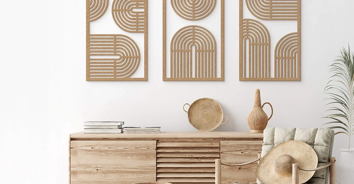 Holzkommode steht an einer weißen Wand mit drei abstrakten Holzbildern darüber. Büchern und Dekorationsgegenständen darauf. Stuhl mit Polster und Strohhut daneben. Pflanze im Hintergrund.