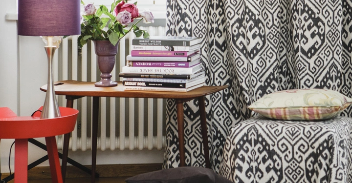 Galerie-Bild 12: Lampe mit violettem Schirm steht auf einem Tisch mit zwei Stapeln Bücher und einer Vase mit rosa Blumen; im Hintergrund ein Heizkörper und Vorhänge mit geometrischem Muster.