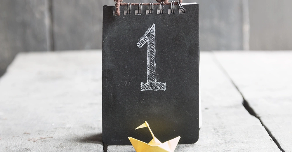 Schwarzes Spiralnotizbuch mit der aufgedruckten weißen Zahl 1 steht auf einem Holztisch; kleines gelbes Papierboot befindet sich davor. Grauer Holzhintergrund.