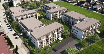 Mehrfamilienhäuser stehen auf einem grünen Rasenbereich mit Bäumen und Gehwegen angeordnet Autos parken auf dem daneben verlaufenden Straßenbereich im Hintergrund sind weitere Gebäude zu sehen.