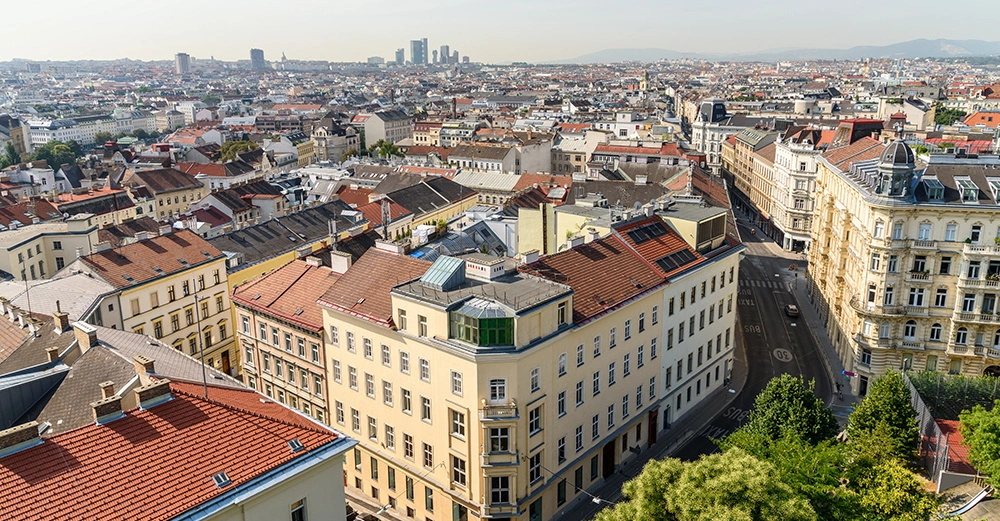 Große Anzahl von Gebäuden mit roten und beigen Dächern verteilen sich in einer Stadtlandschaft mit vereinzelten Hochhäusern im Hintergrund und Bäumen im Vordergrund bei hellem Tageslicht.