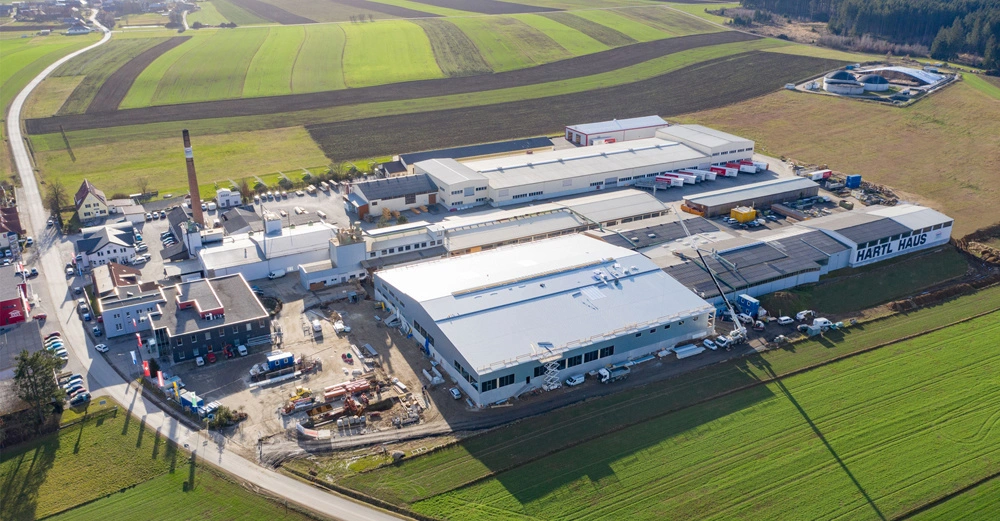 Großes industrielles Gebäude mit Aufschriften HARTL HAUS und DIENST WERK auf den Gebäuden Autos und Baumaterialien umgeben die Anlage umliegende grüne und braune Felder Straße verläuft durch benachbarte Häuserlandschaft
