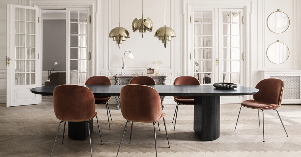 Ovaler schwarzer Esstisch stehend in einem eleganten weißen Raum umgeben von sechs braunen Stühlen drei hängenden goldenen Lampen und hohen Doppelflügeltüren im Hintergrund dekorative Wanddetails