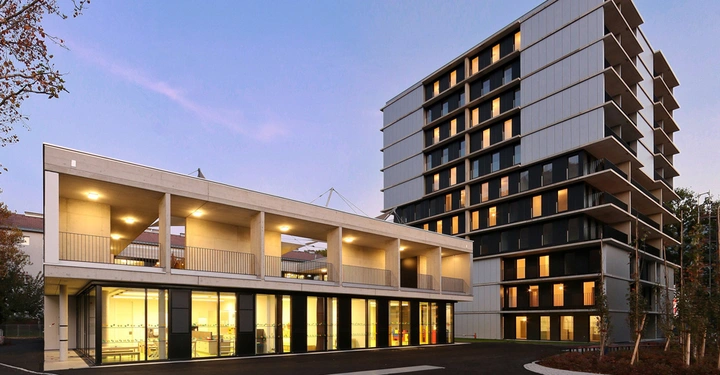 Galerie-Bild 9: Ein modernes zweistöckiges Gebäude mit großen Glasfenstern im Erdgeschoss steht neben einem höheren Wohnhochhaus in einer städtischen Umgebung bei Dämmerung.