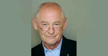 Ein älterer Mann mit kurzen grauen Haaren und Brille lächelt leicht in die Kamera, trägt ein blaues Hemd und einen schwarzen Blazer, Hintergrund unscharf grauer Farbton.